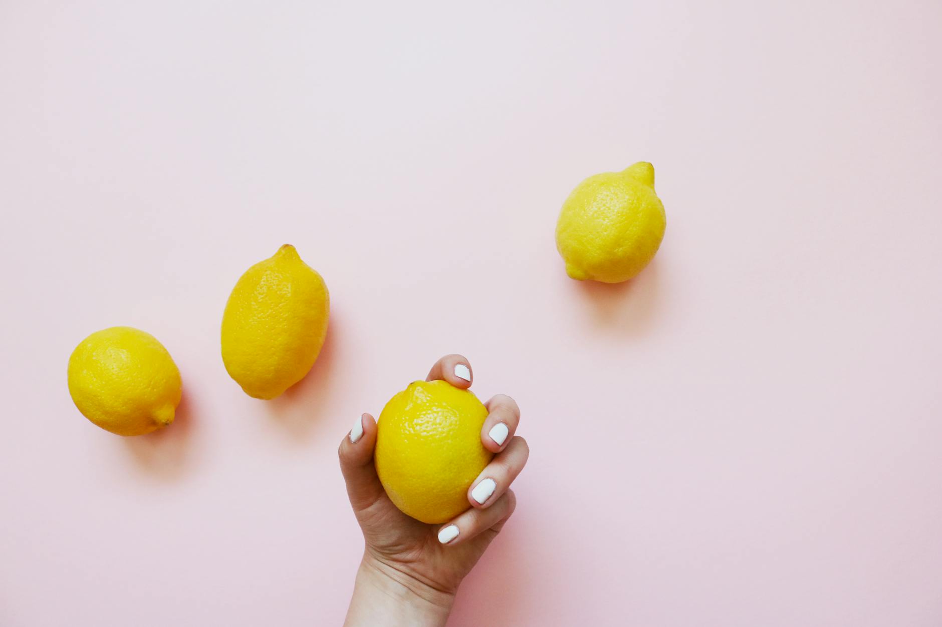 A hand holding a bright yellow lemon on a soft pink background, symbolizing fresh pleasure and intimacy.