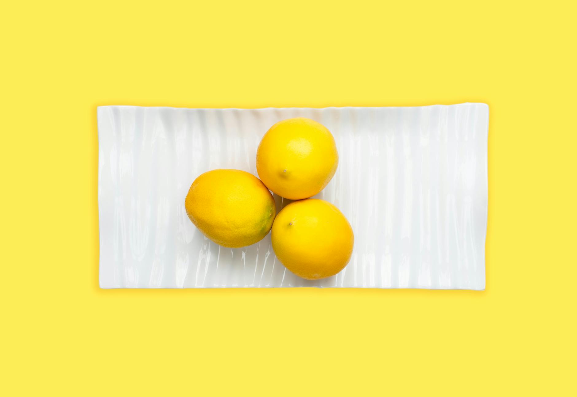 Three fresh lemons arranged on a white plate with a vibrant yellow background, emphasizing freshness and citrus appeal.