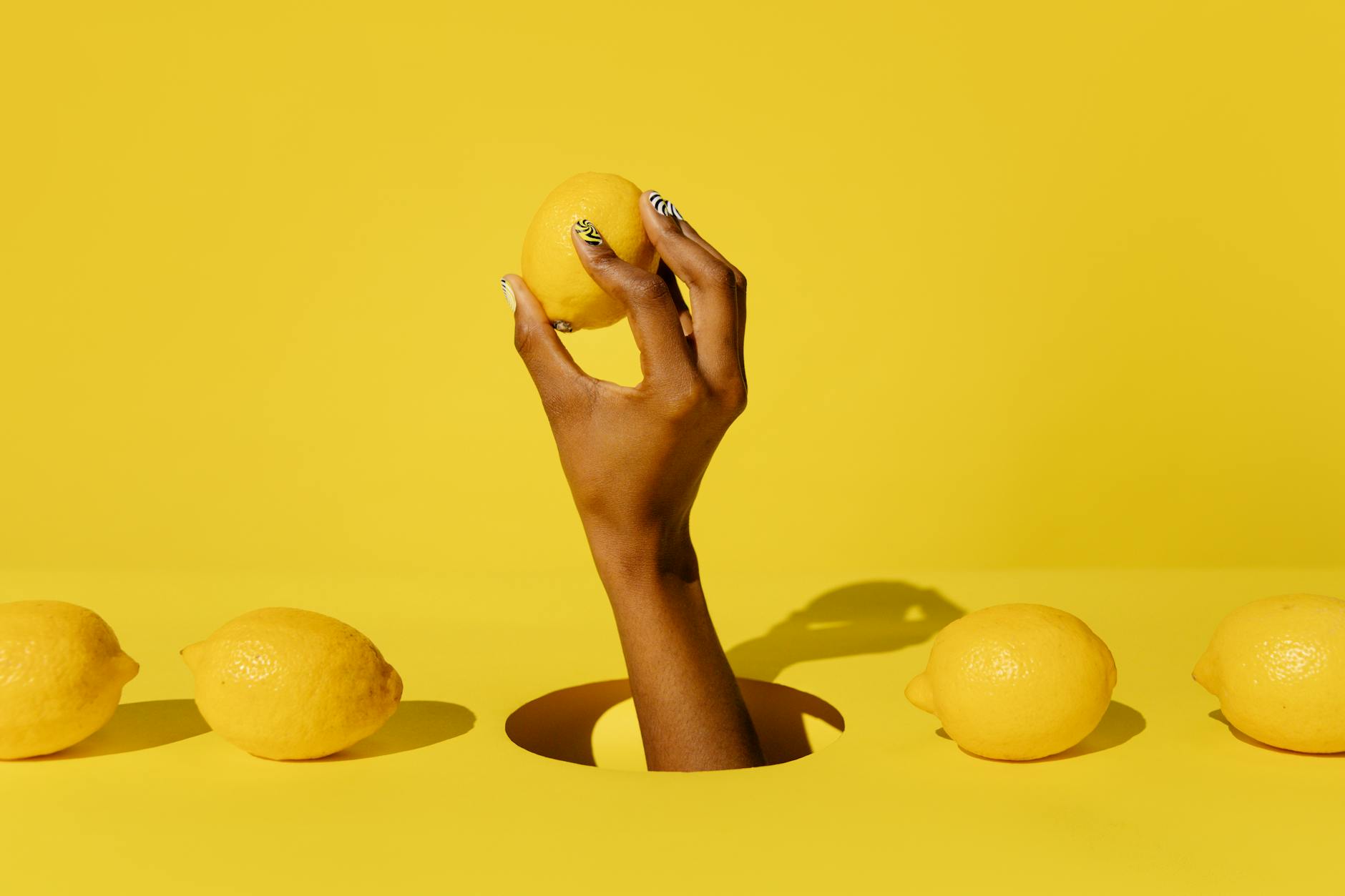 A hand holding a fresh lemon against a bright yellow background, symbolizing the citrusy energy and renewed pleasure from Lemon Clit Massager