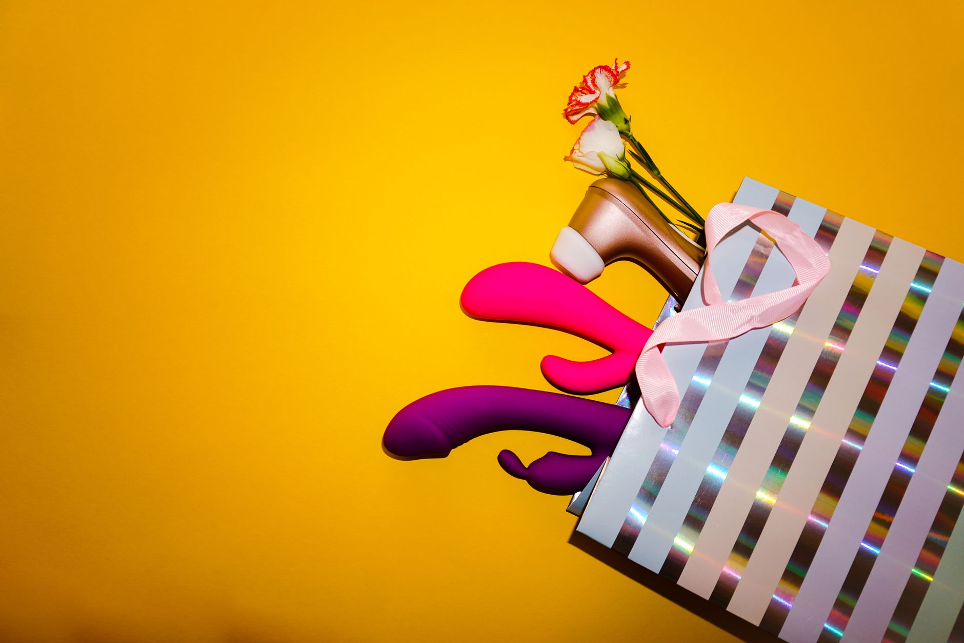 Colorful vibrators including lemon clitoral vibrators displayed with flowers in a holographic gift bag on a bold yellow background.